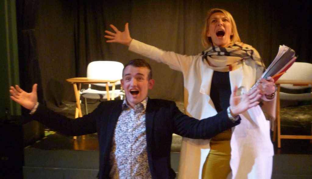 Two performers onstage as part of a play, with their arms raised and mouths open as if they have been making an important announcement